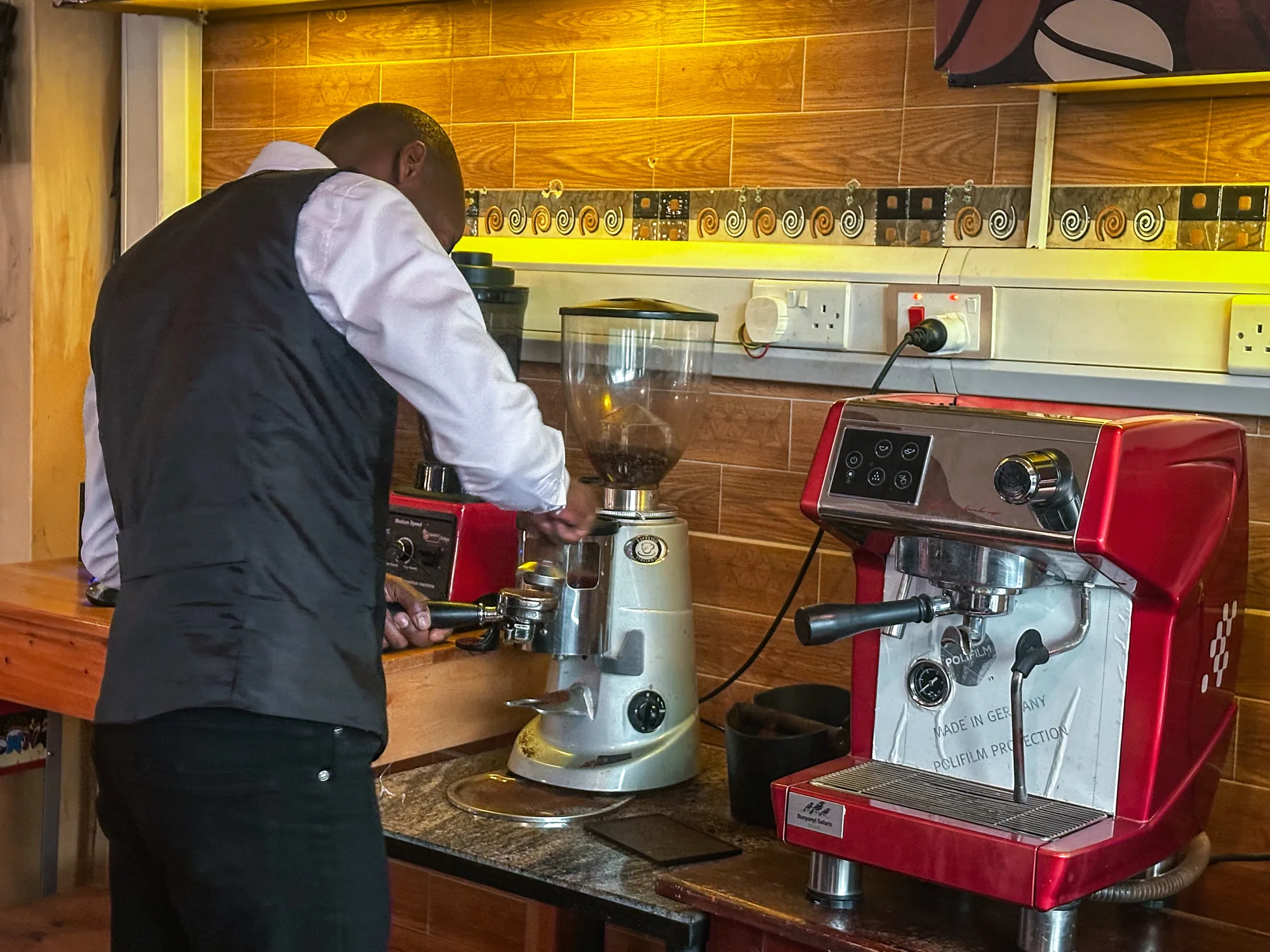 A waistcoated barista grinding coffee at the machine behind the yellow-tiled counter
