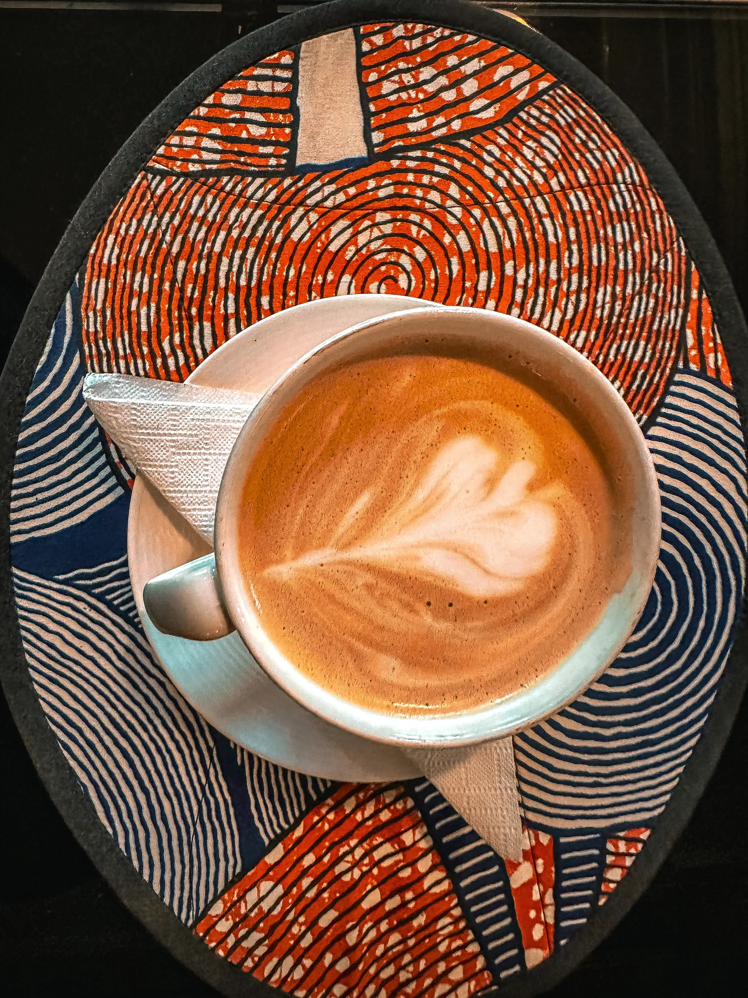 A cappuccino with leaf latte art on a patterned woven mat, seen from above