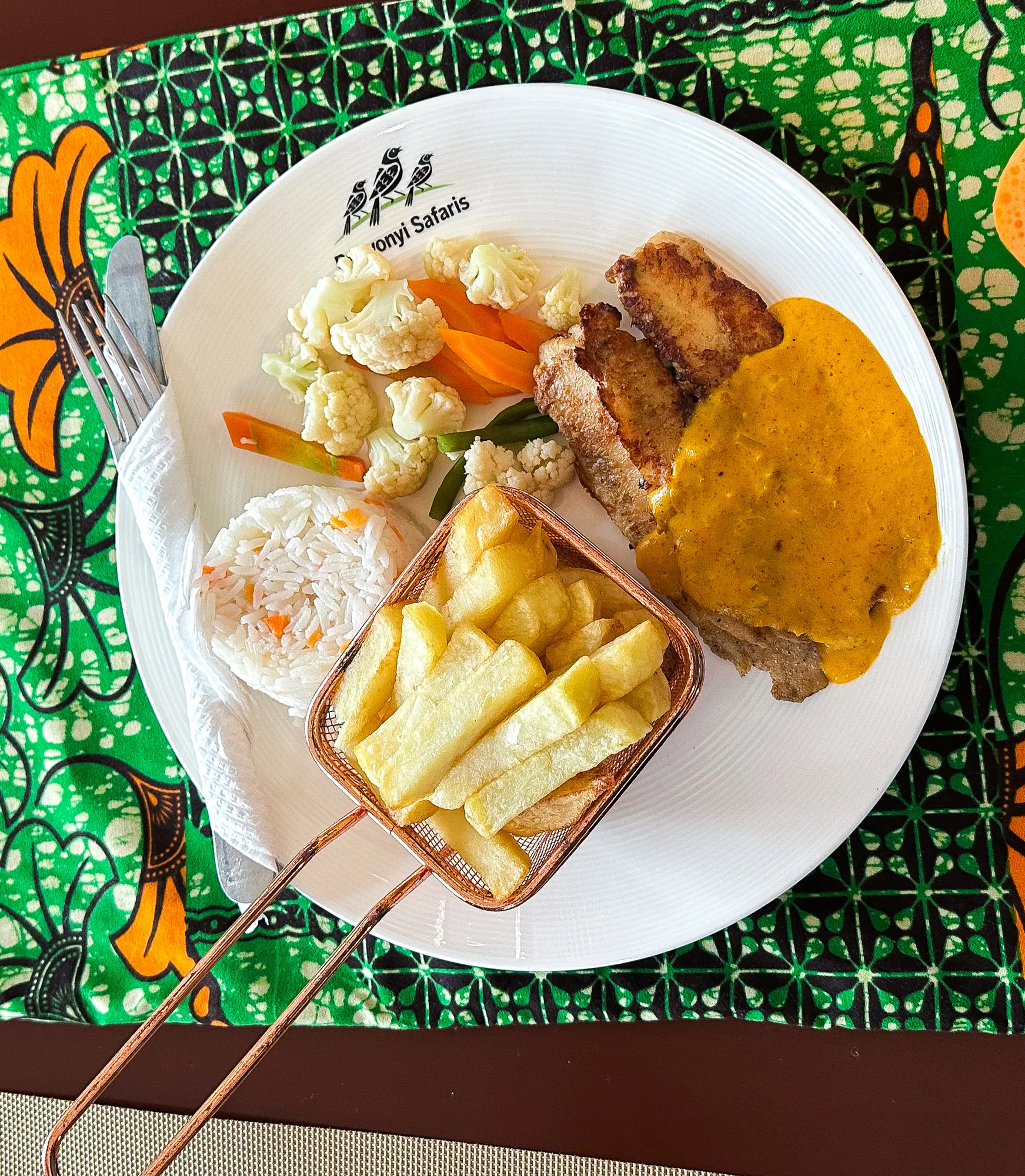 A plated main course of chips, rice, vegetables and meat with sauce on a branded plate