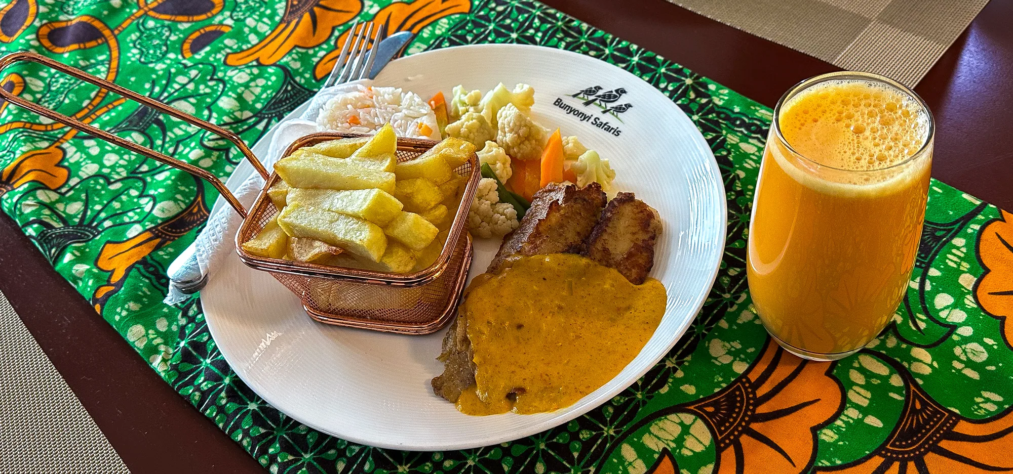 A plate of chips and grilled meat with a glass of passion juice on a green printed cloth