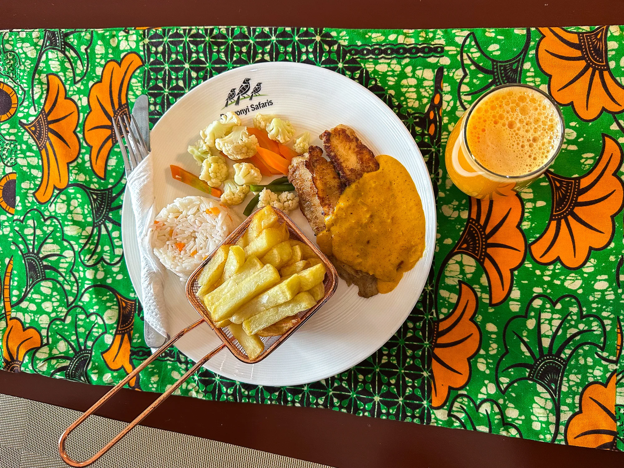 A plate of chips, rice, mixed vegetables and meat beside a glass of juice on an orange printed cloth