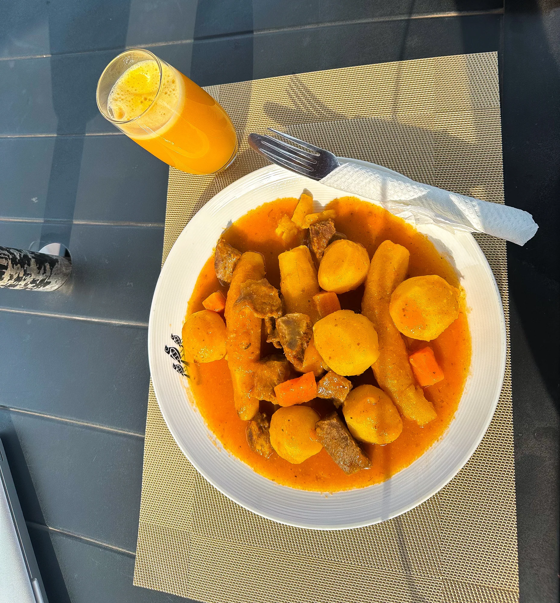 A bowl of beef and potato stew with a glass of juice on a grey table, seen from above
