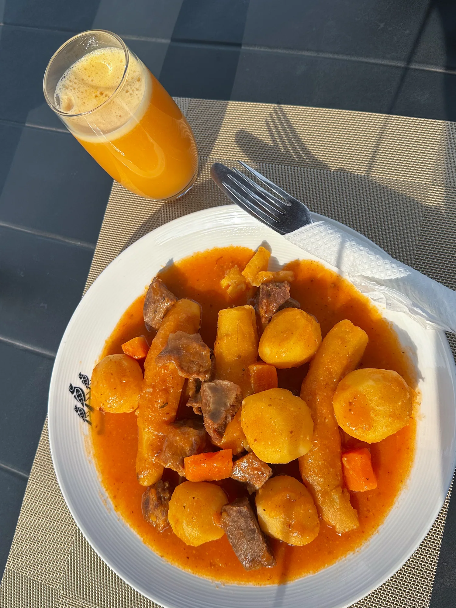A bowl of beef stew with potatoes and carrots next to a glass of juice and cutlery