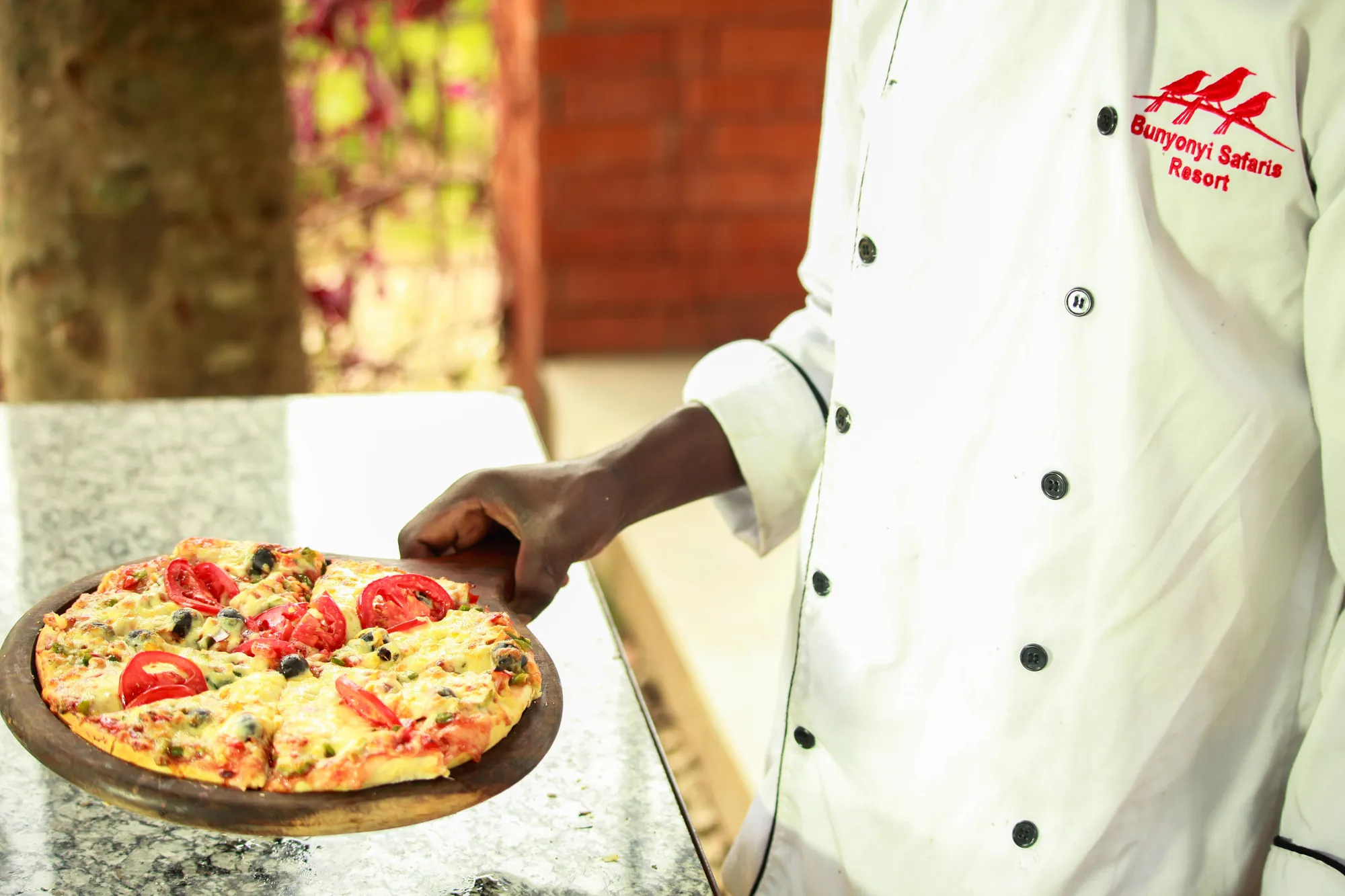 A chef in a branded white jacket lifting a freshly baked pizza on a wooden board
