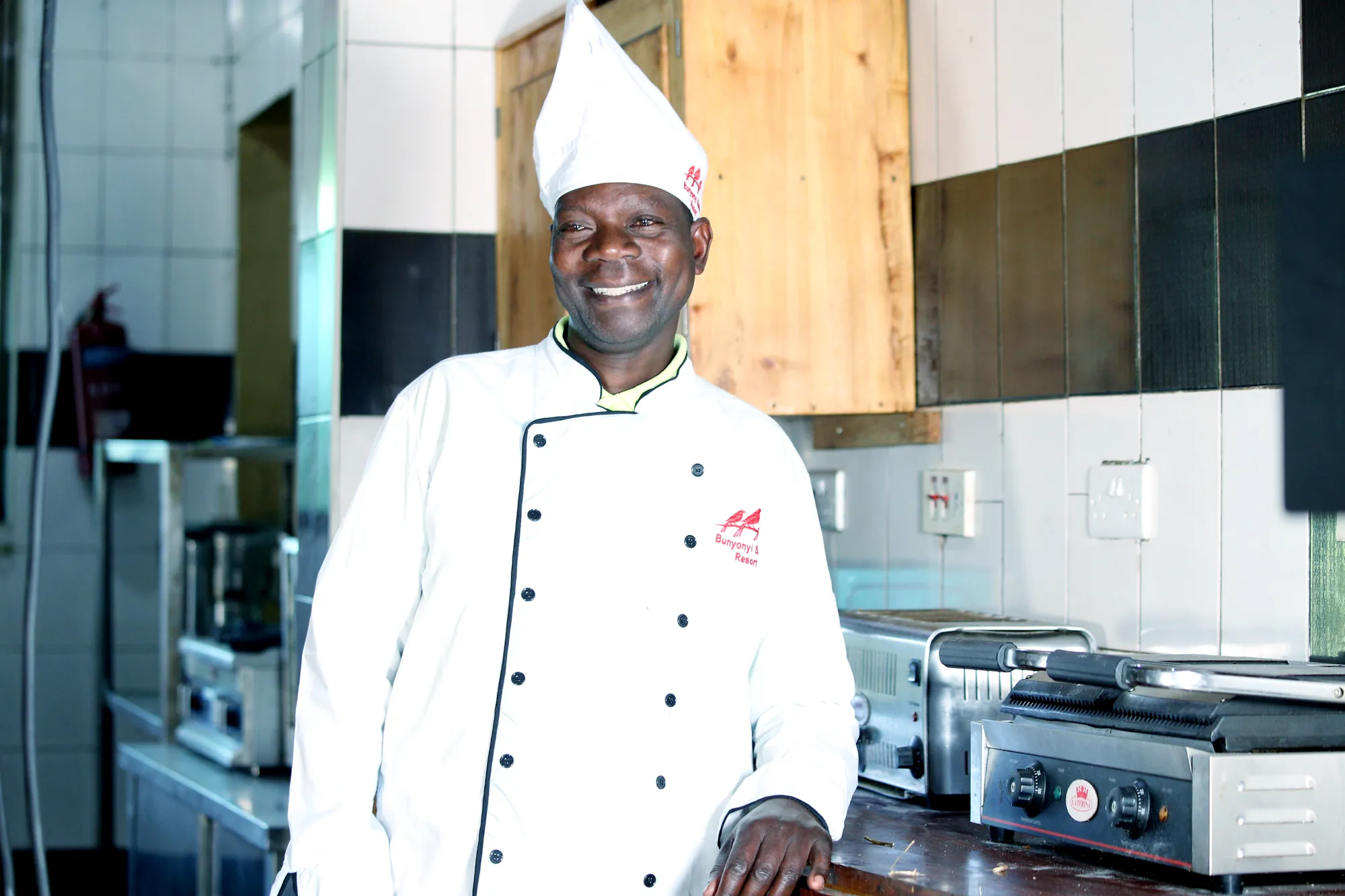 A smiling chef in a tall white hat beside the kitchen range