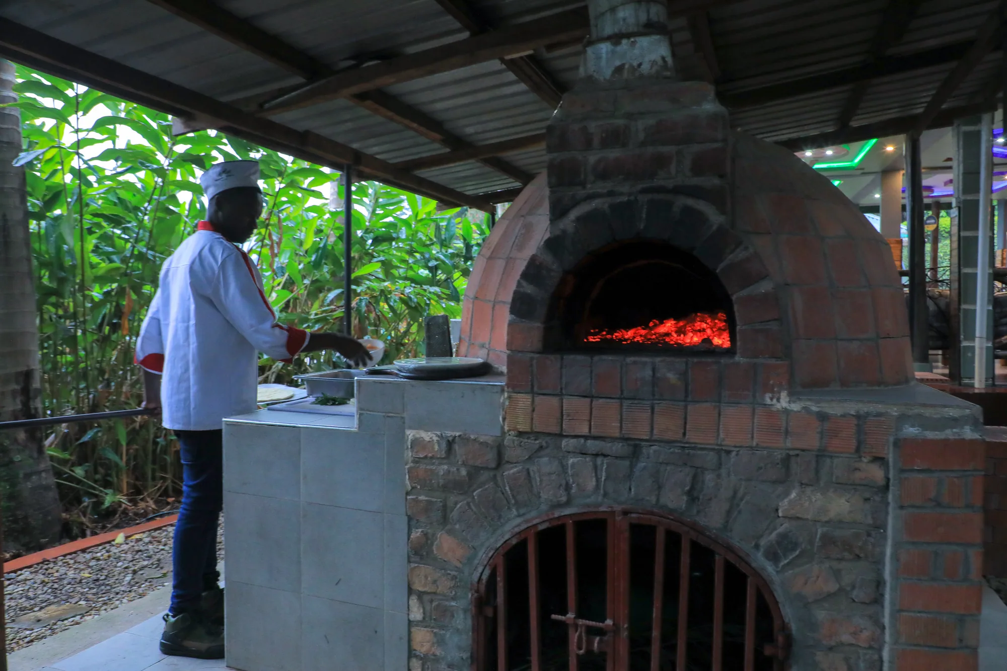 The pizza oven with its stone base and chimney, a cook working at the peel beside it