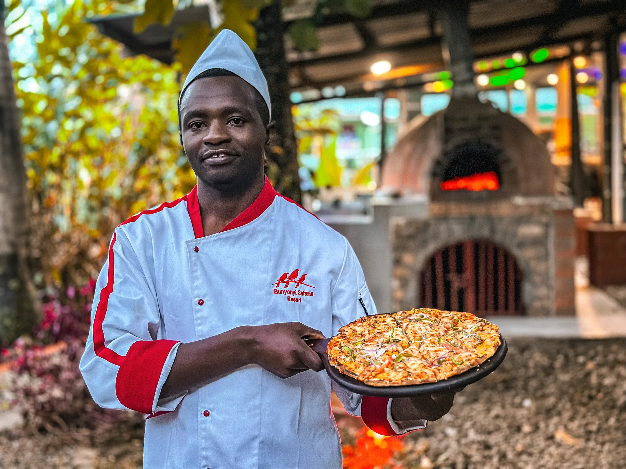 A chef holding up a finished pizza on a metal tray in front of the lit pizza oven