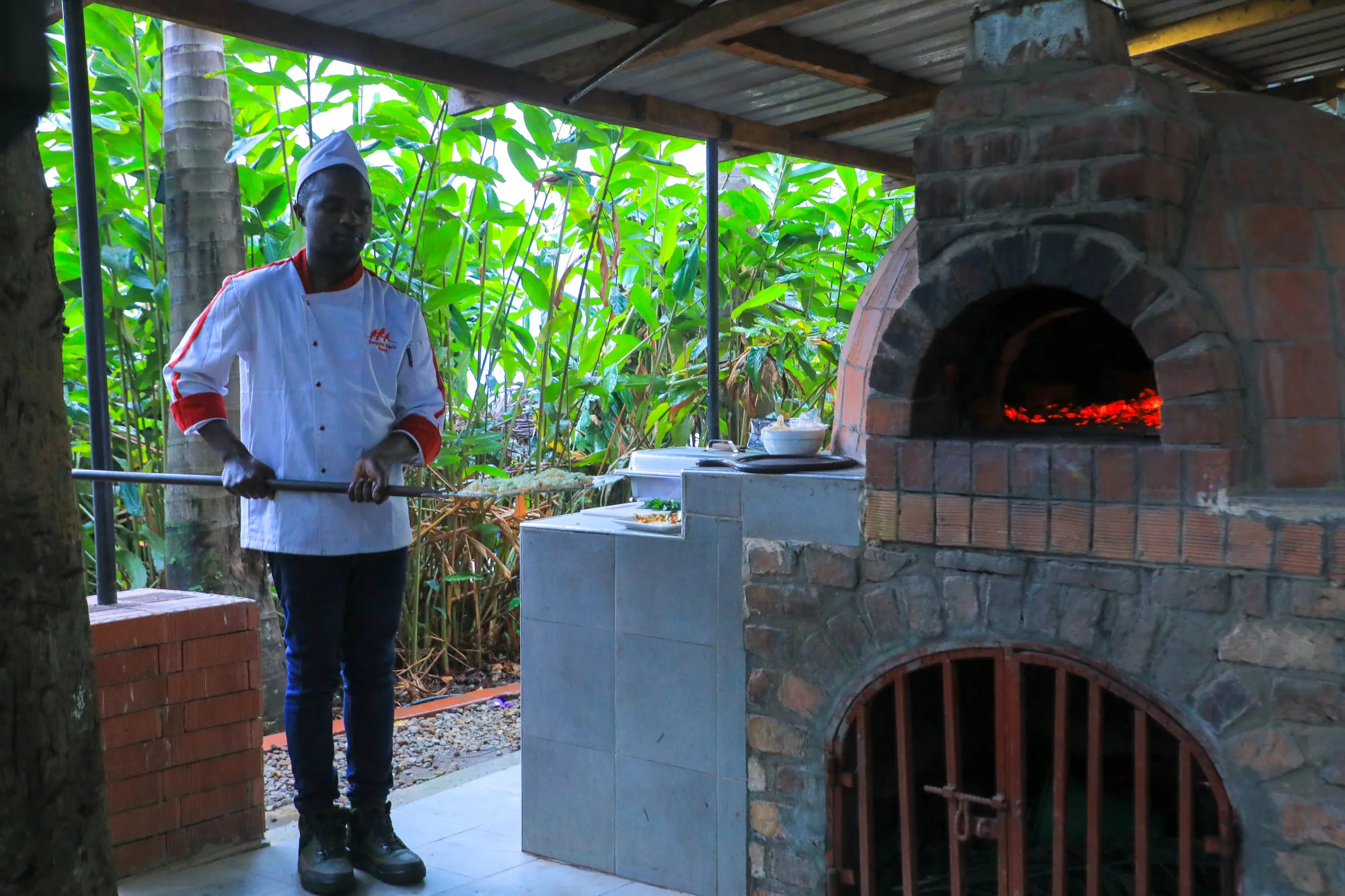A cook standing with a tray beside the pizza oven, the fire glowing inside