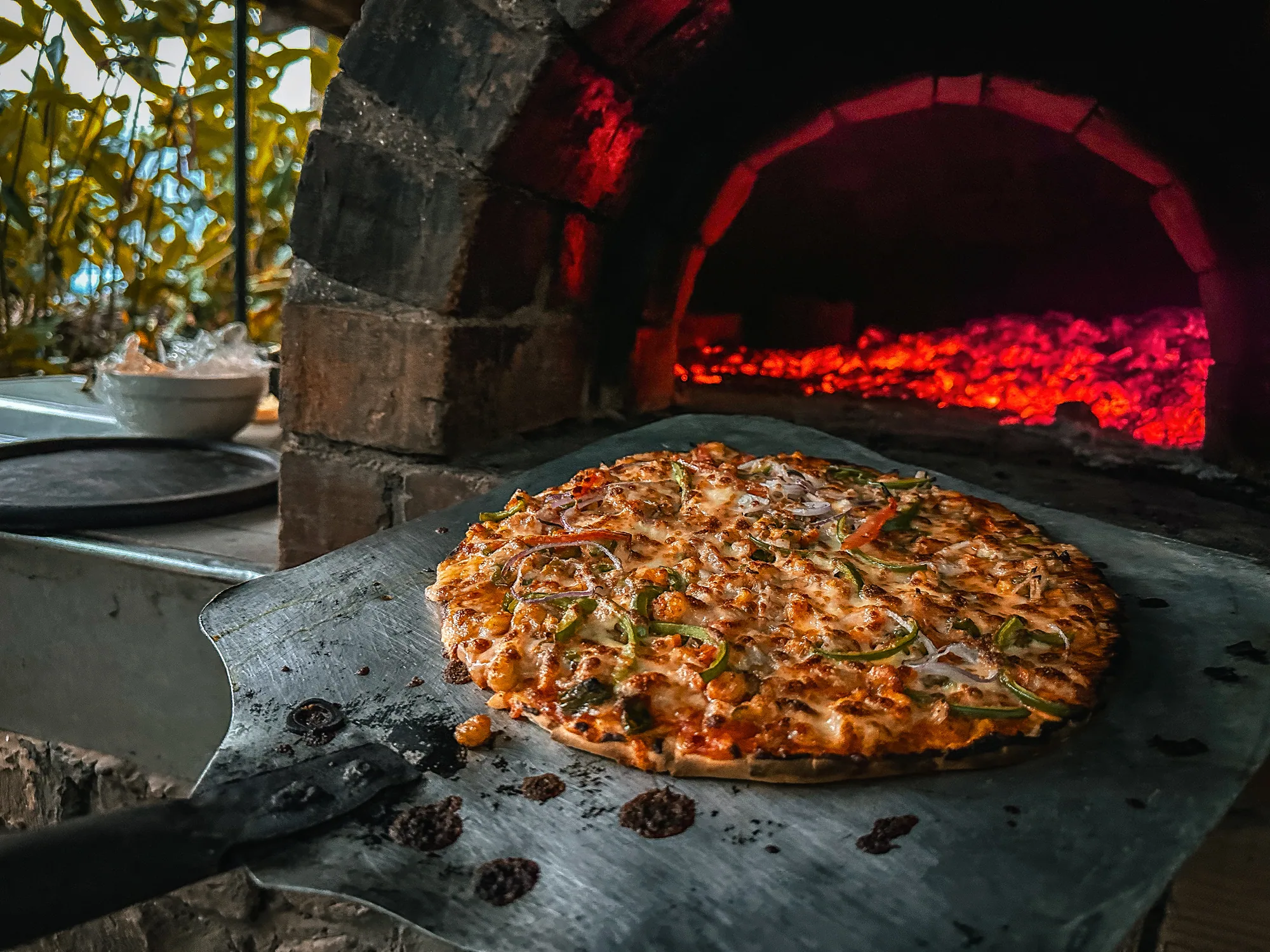 A pizza on a metal tray in front of the glowing embers inside the wood-fired oven