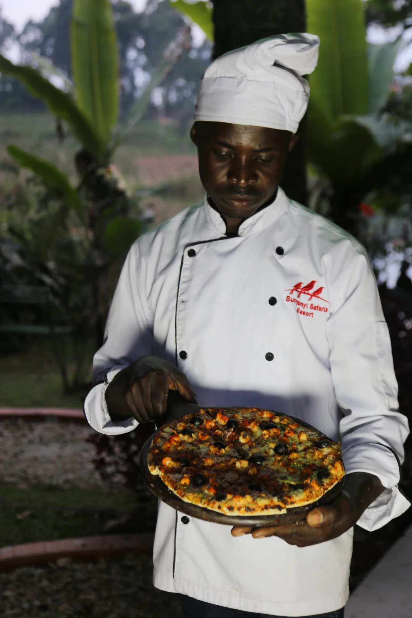 A cook in a white hat holding a pizza on a tray in the garden by the oven