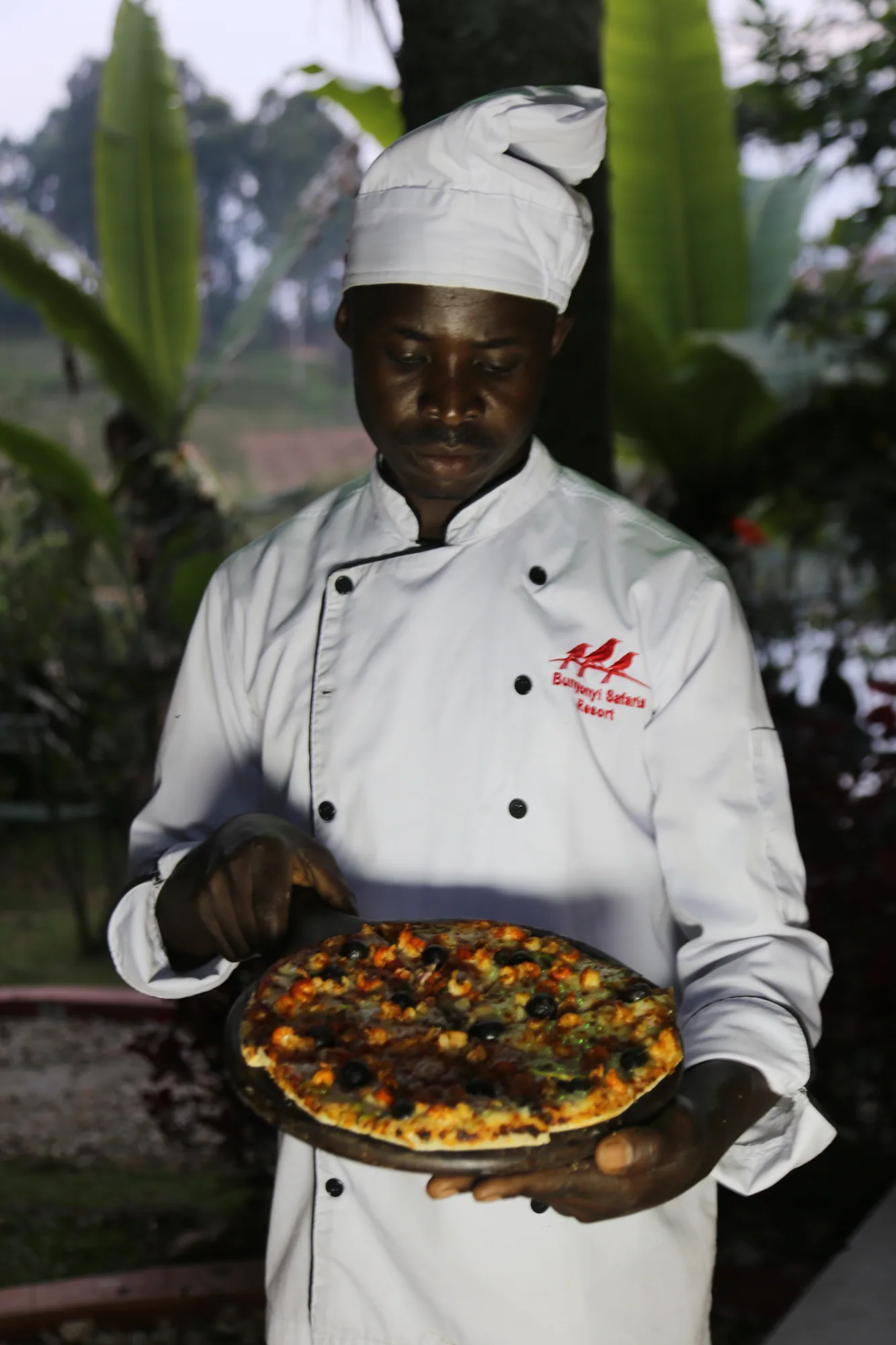 A cook holding a pizza on a tray with banana leaves behind