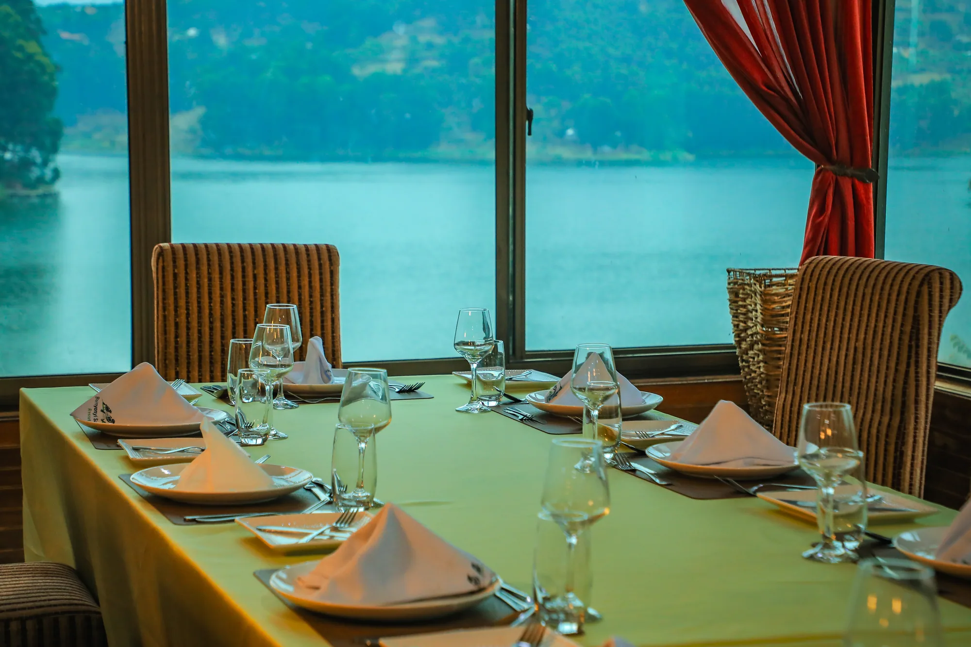 A long dining table laid with glassware and folded napkins in front of a window over the lake