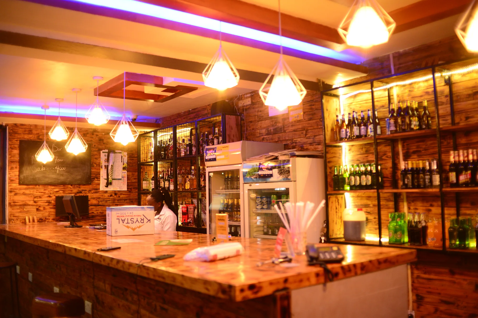 The lit bar counter with pendant lamps, bottle shelves and a drinks fridge