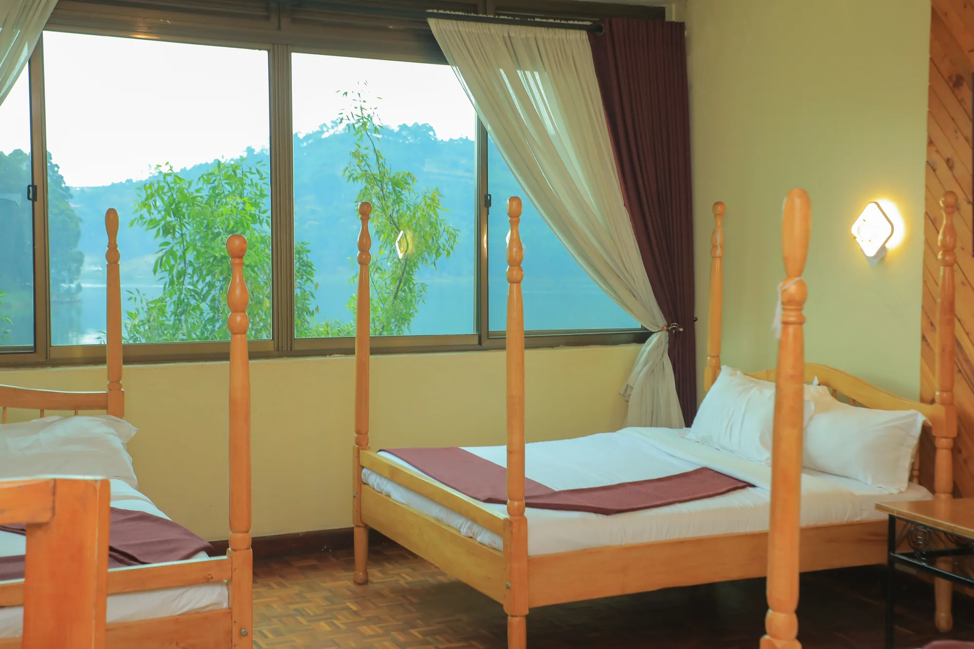 A four-poster wooden bed with white linen beside a wide window over the hills