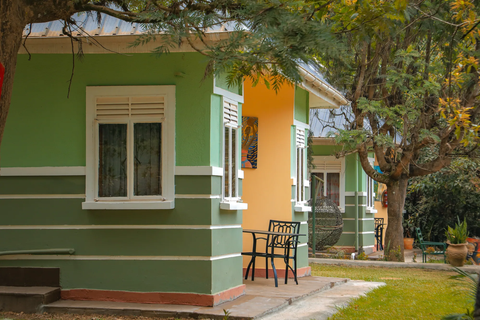 The corner of a green cottage with white-framed windows and chairs outside under a tree