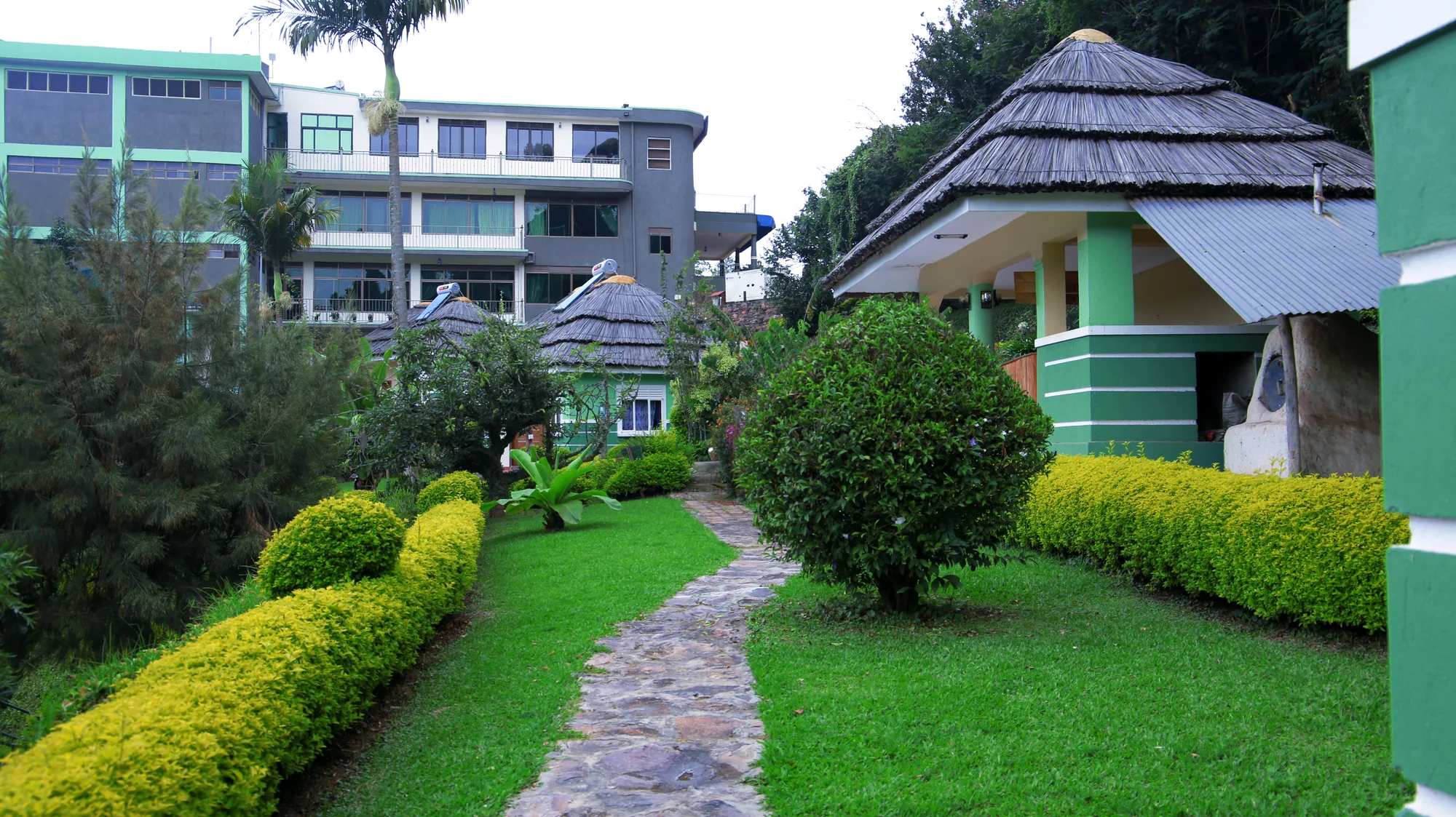 A paved garden path between clipped hedges leading past thatched cottages to the main hotel building