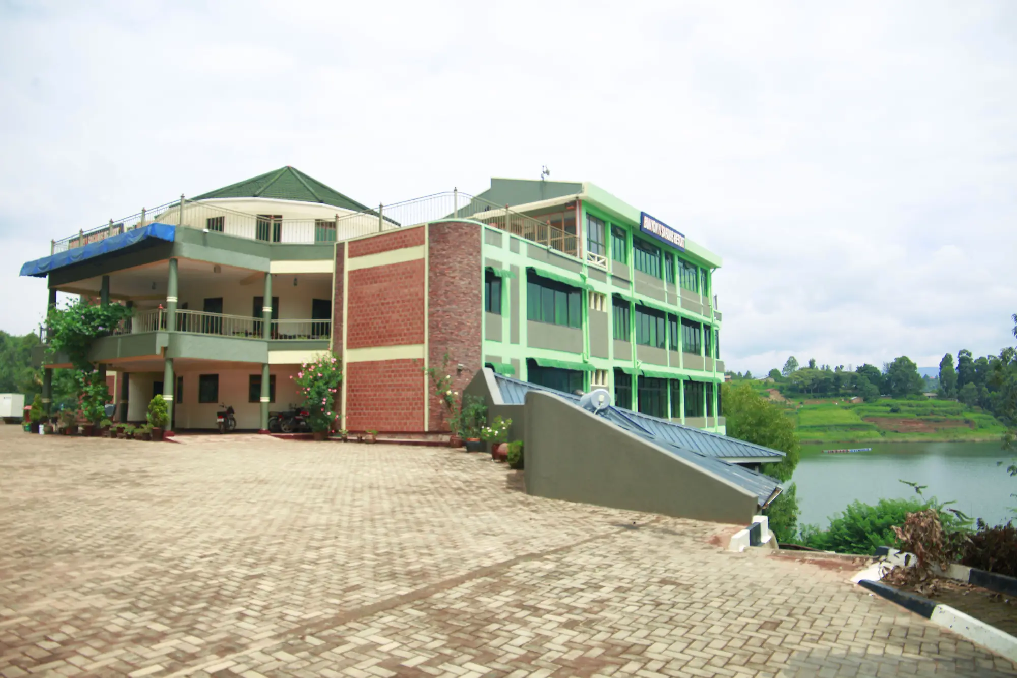 Green and brick multi-storey building with a paved forecourt beside the lake