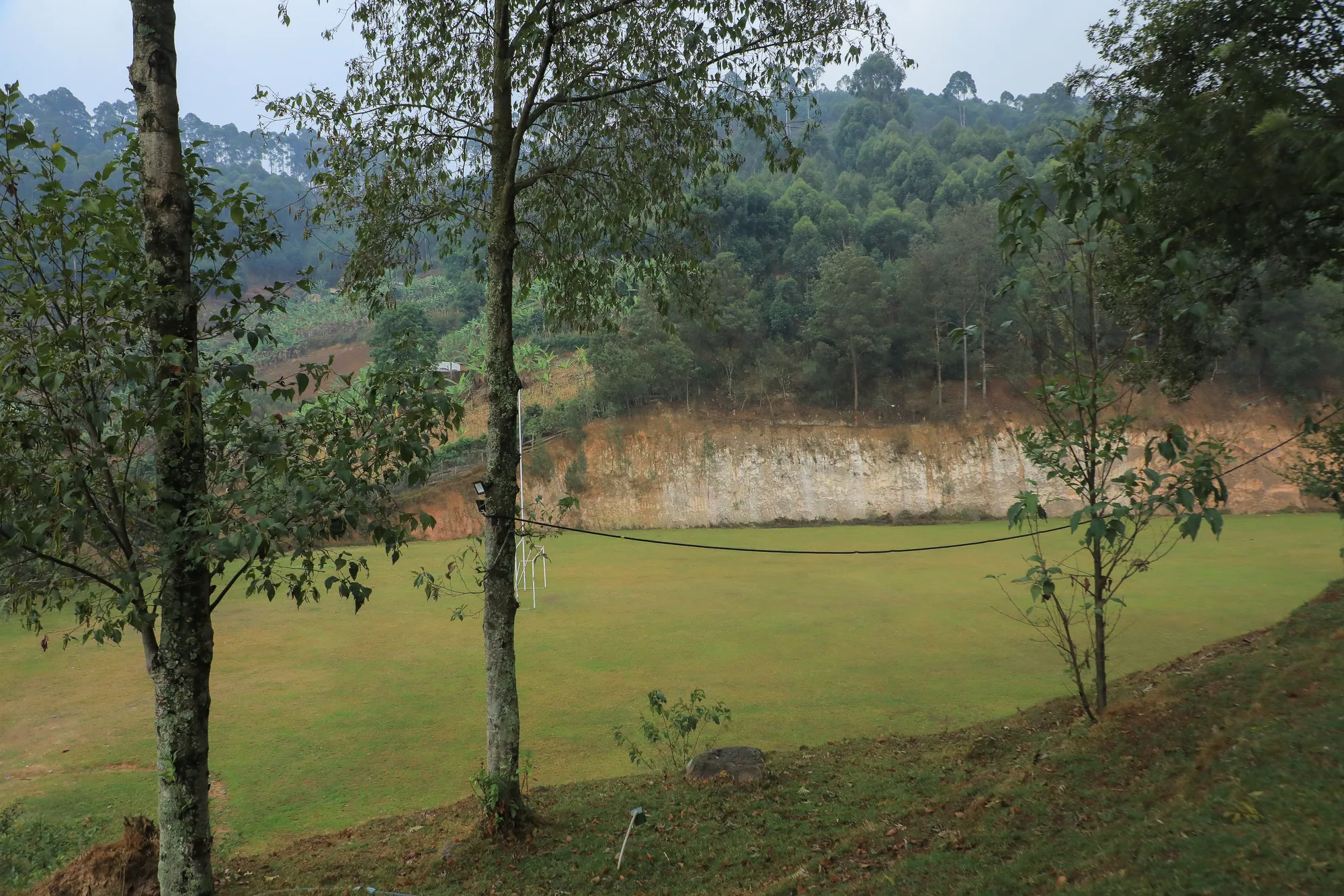 An open grass field on the hilltop site with young trees and a red earth bank