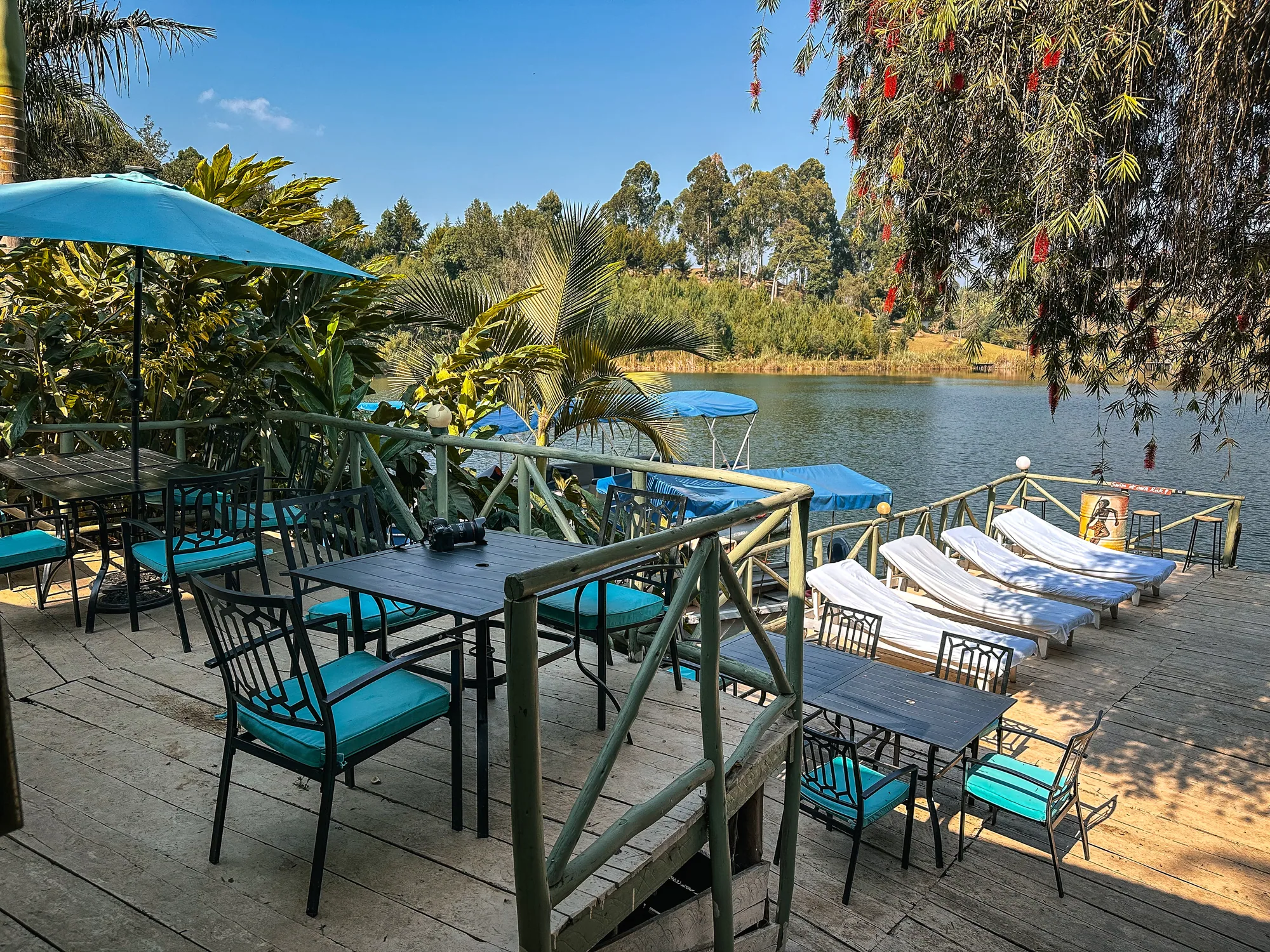Deck tables under turquoise parasols with loungers behind and palms at the lakeside