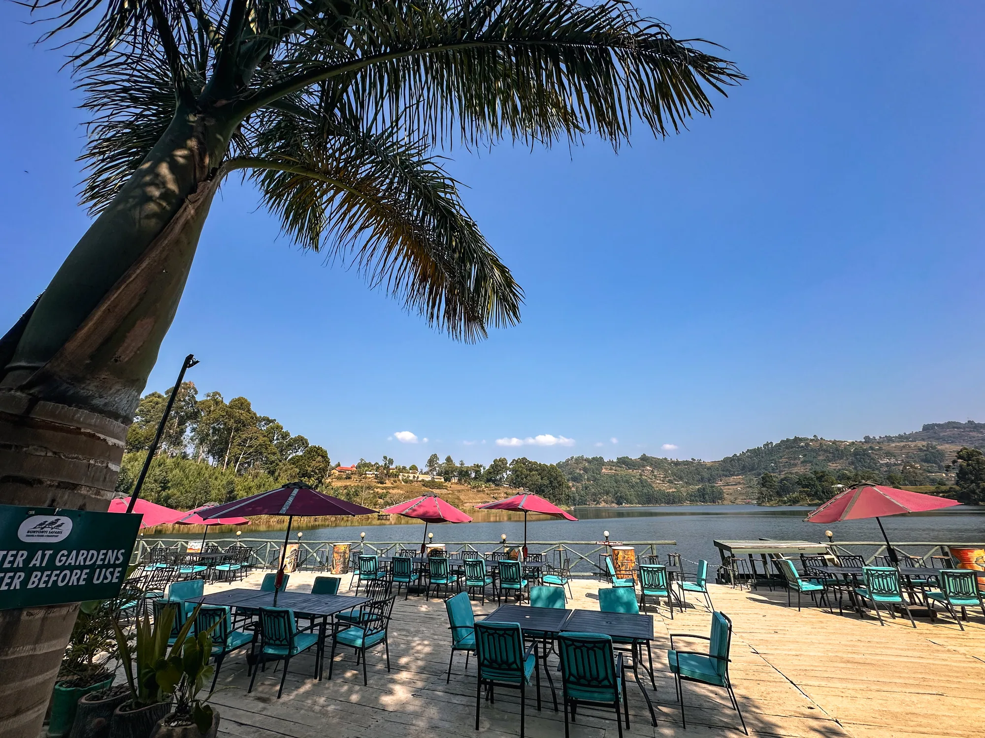 Deck seating under parasols framed by a palm, with the lake behind and a garden sign in front