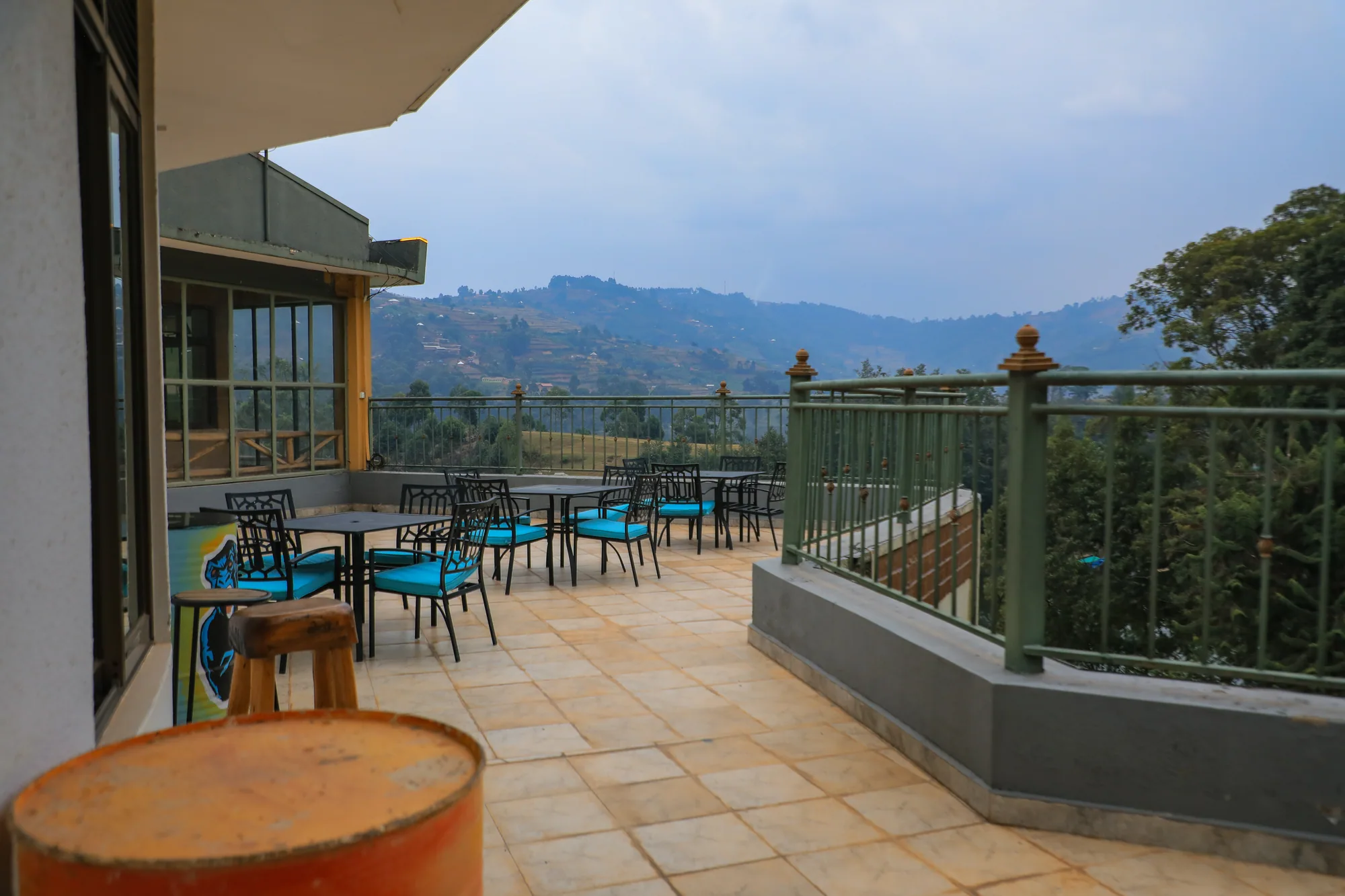 A rooftop terrace of black tables and turquoise chairs enclosed by a metal railing, hills beyond