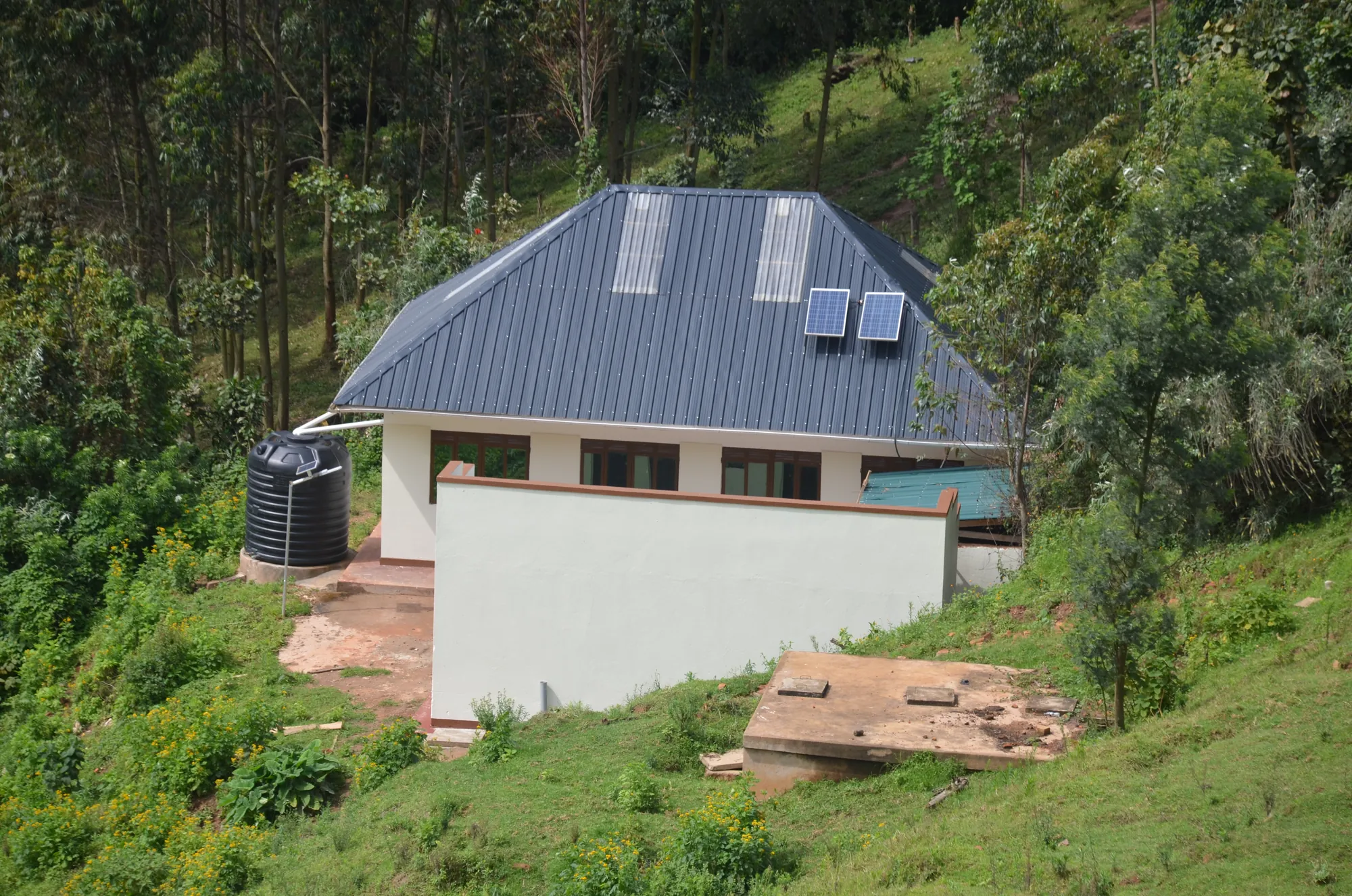 A white building with solar panels on its roof and a water tank at the side on the hillside