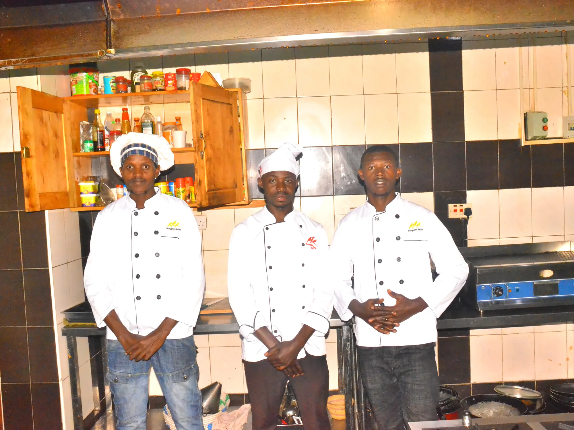 Three trainee cooks in whites standing in front of the kitchen shelves and range