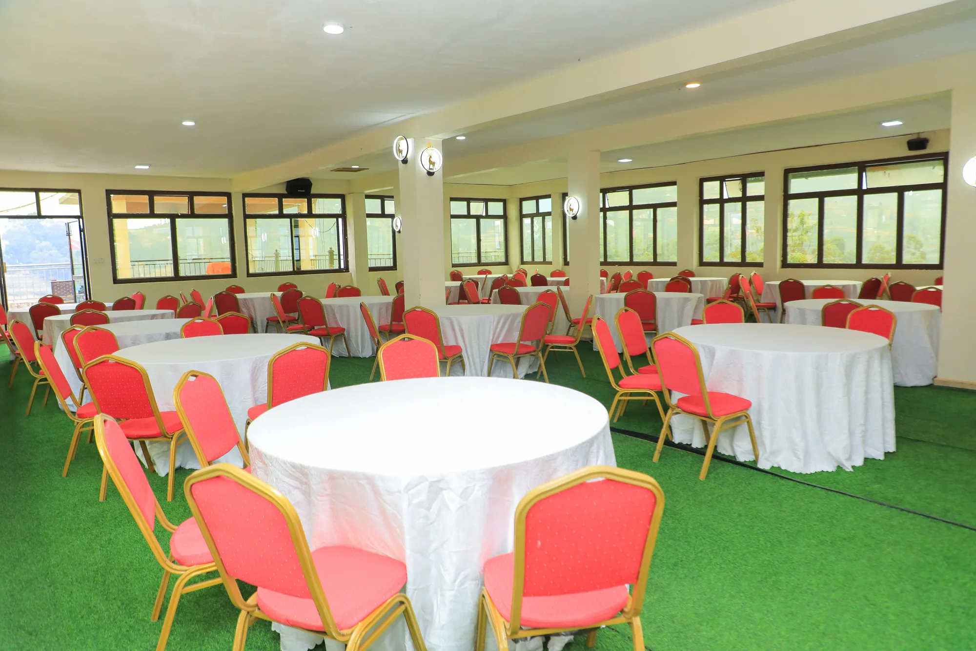 A banqueting hall set with round tables, white cloths and red chairs on green carpet