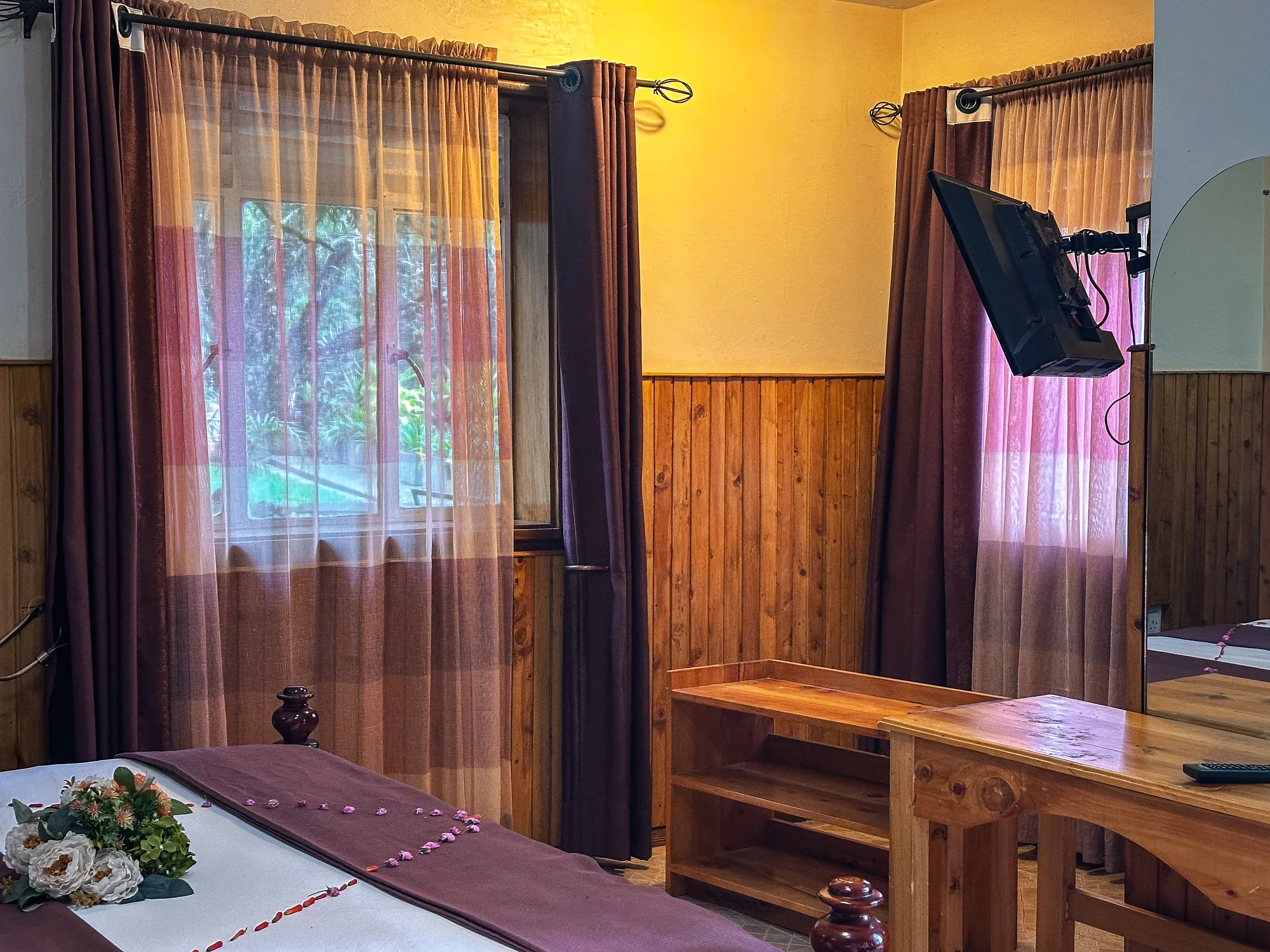 A timber-panelled room with a bed dressed in a red runner and petals beside curtained windows