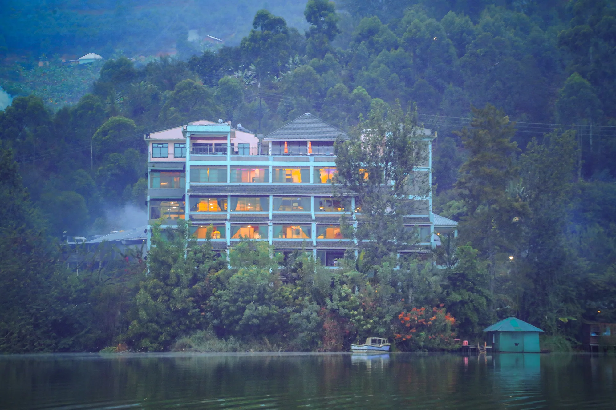 The hotel block with its windows lit, seen across the lake through morning haze