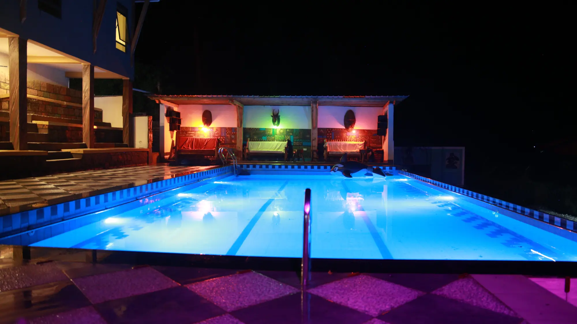 Outdoor swimming pool lit blue at night with coloured lights on the pool house