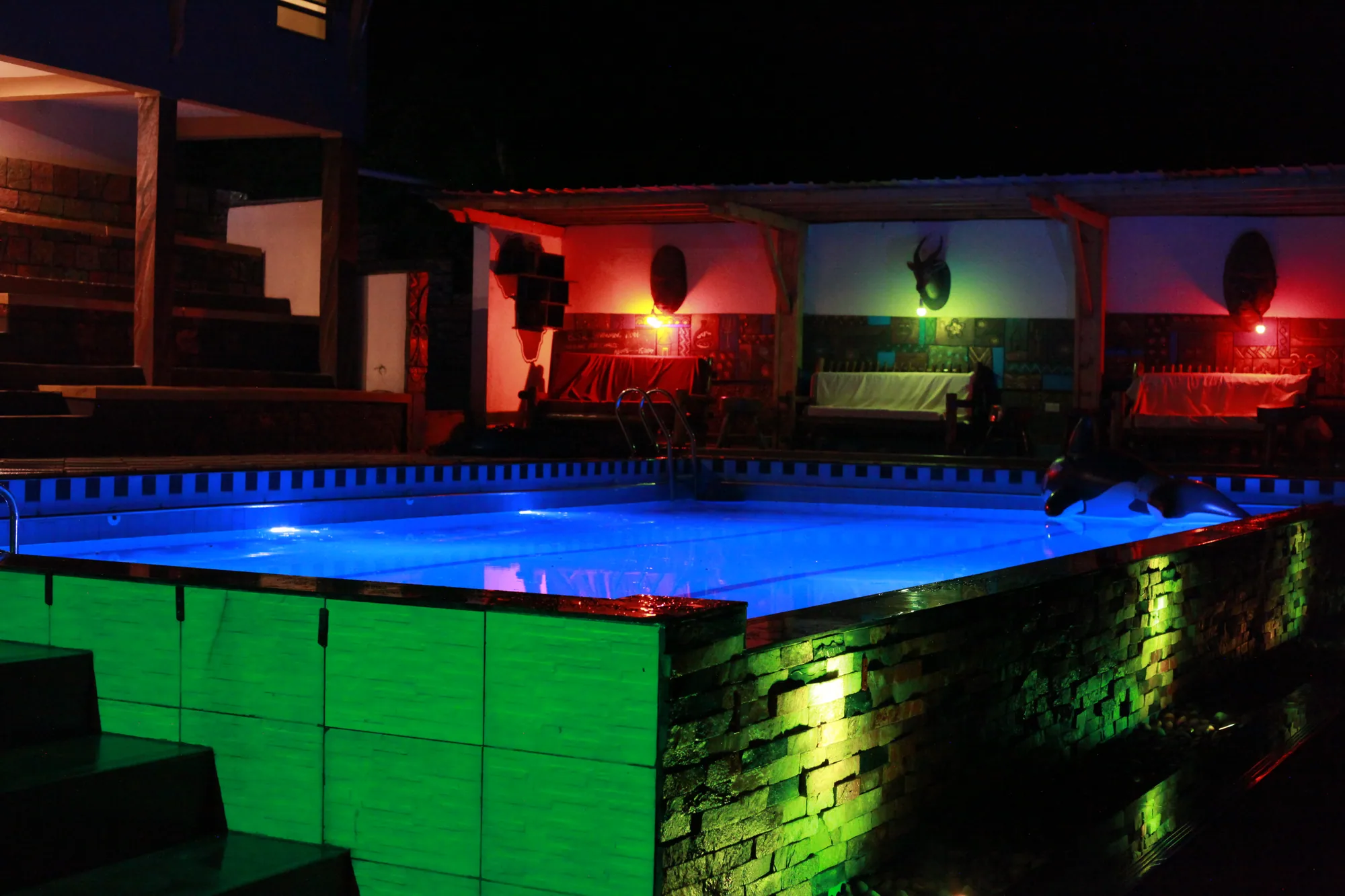 The swimming pool lit blue at night with red and green lighting on the terrace behind