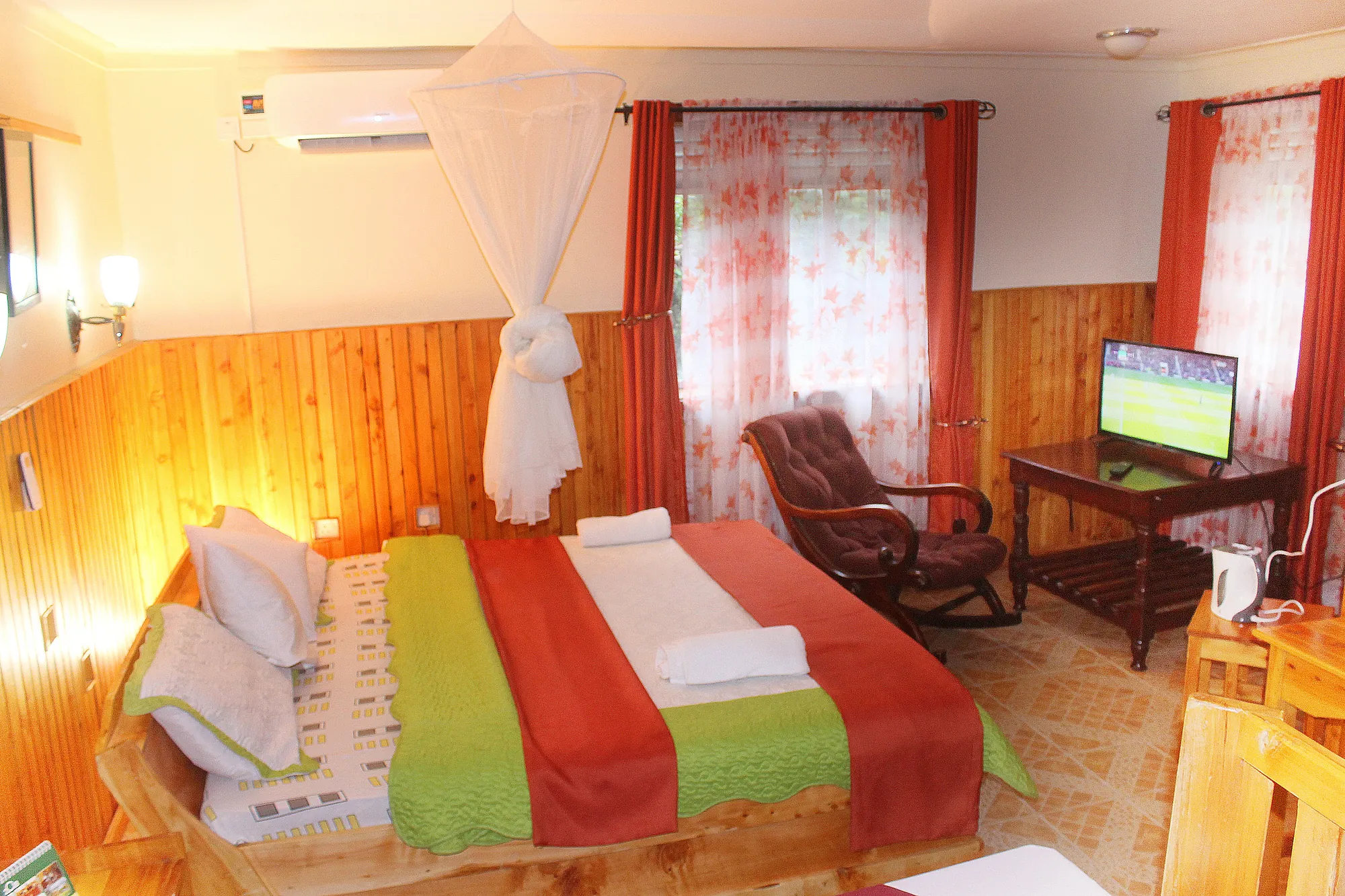 A cottage bedroom with timber panelling, a red bed runner, mosquito net and a television