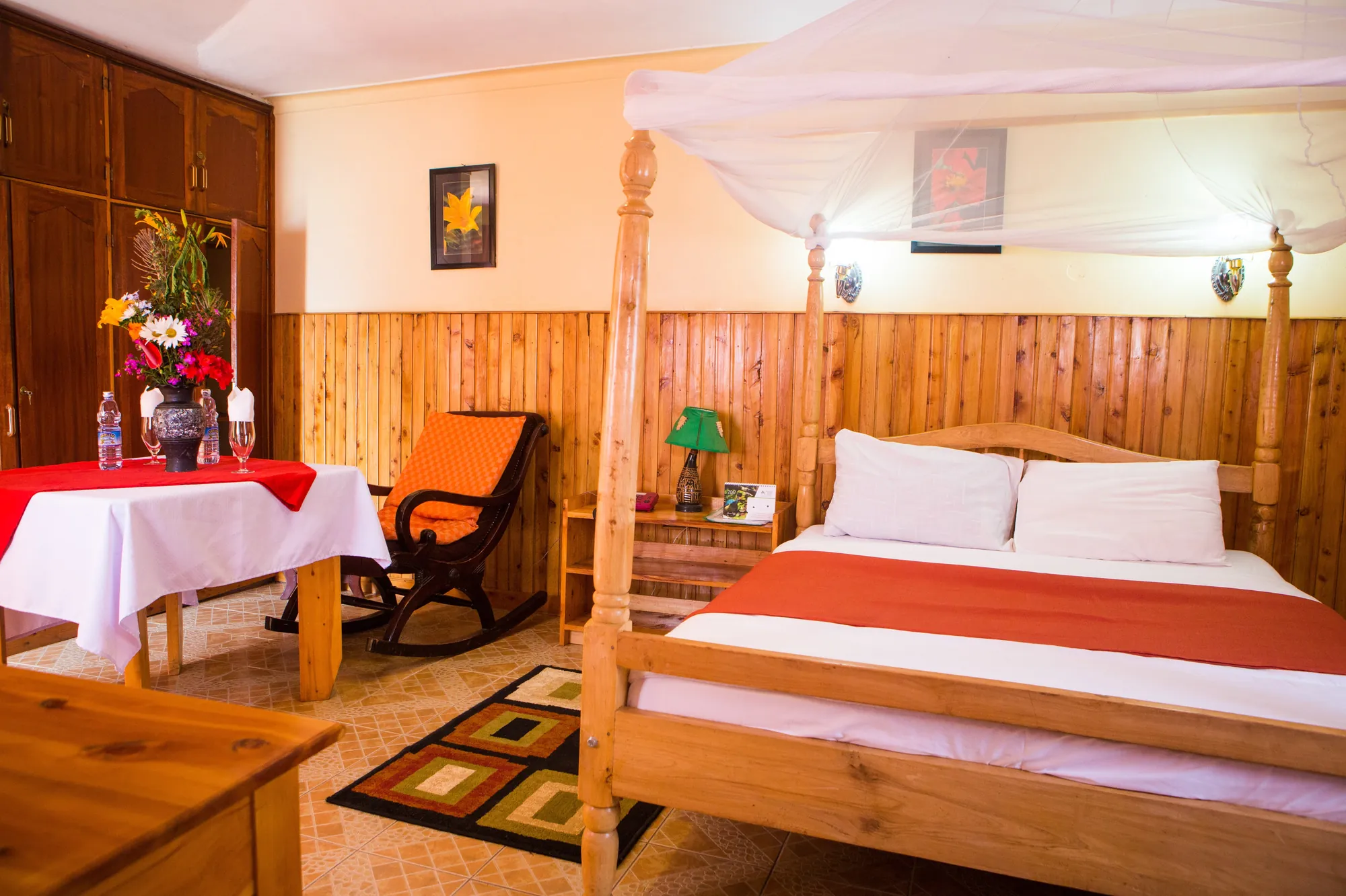 A four-poster bed with a red runner beside a laid table and a rocking chair in a cottage room