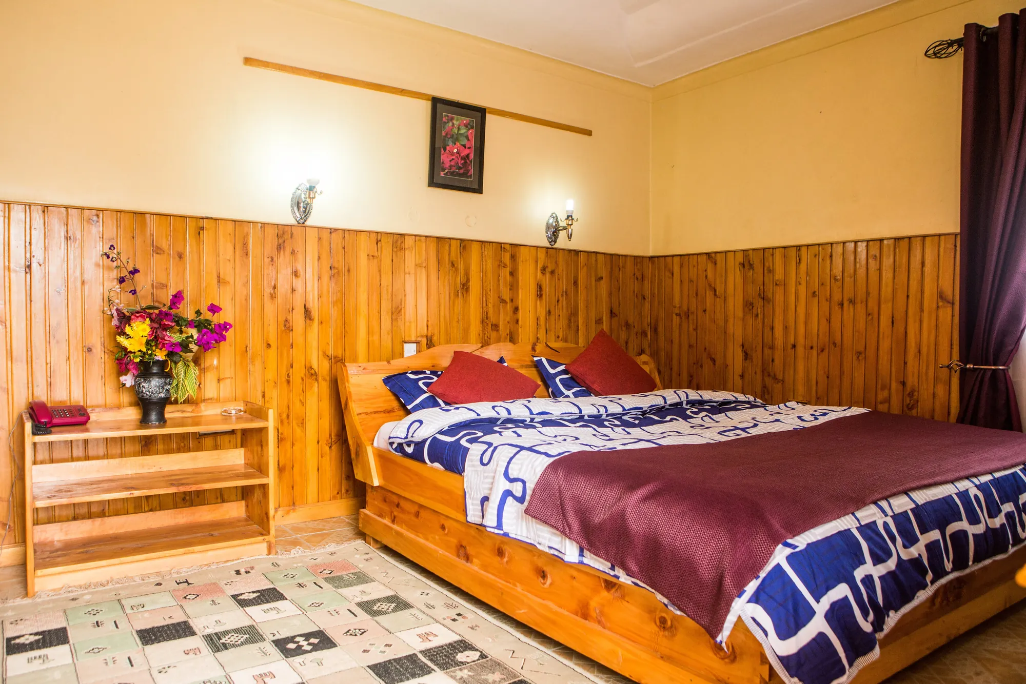 Double / Twin Room: double bed with a patterned cover and red cushions beside a vase of flowers