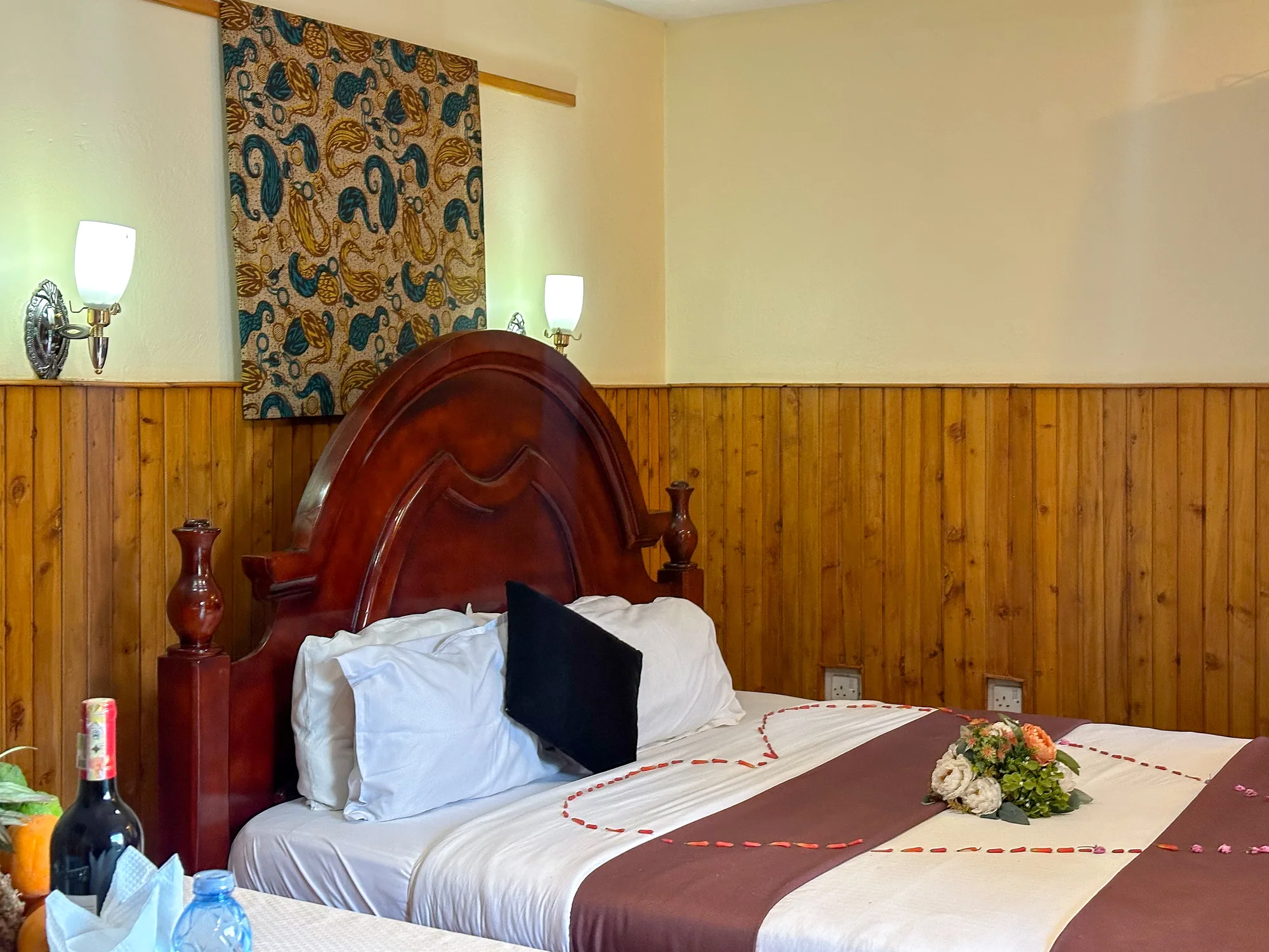Cottage: carved wooden bed with a maroon runner and a flower posy, welcome drinks at the bedside