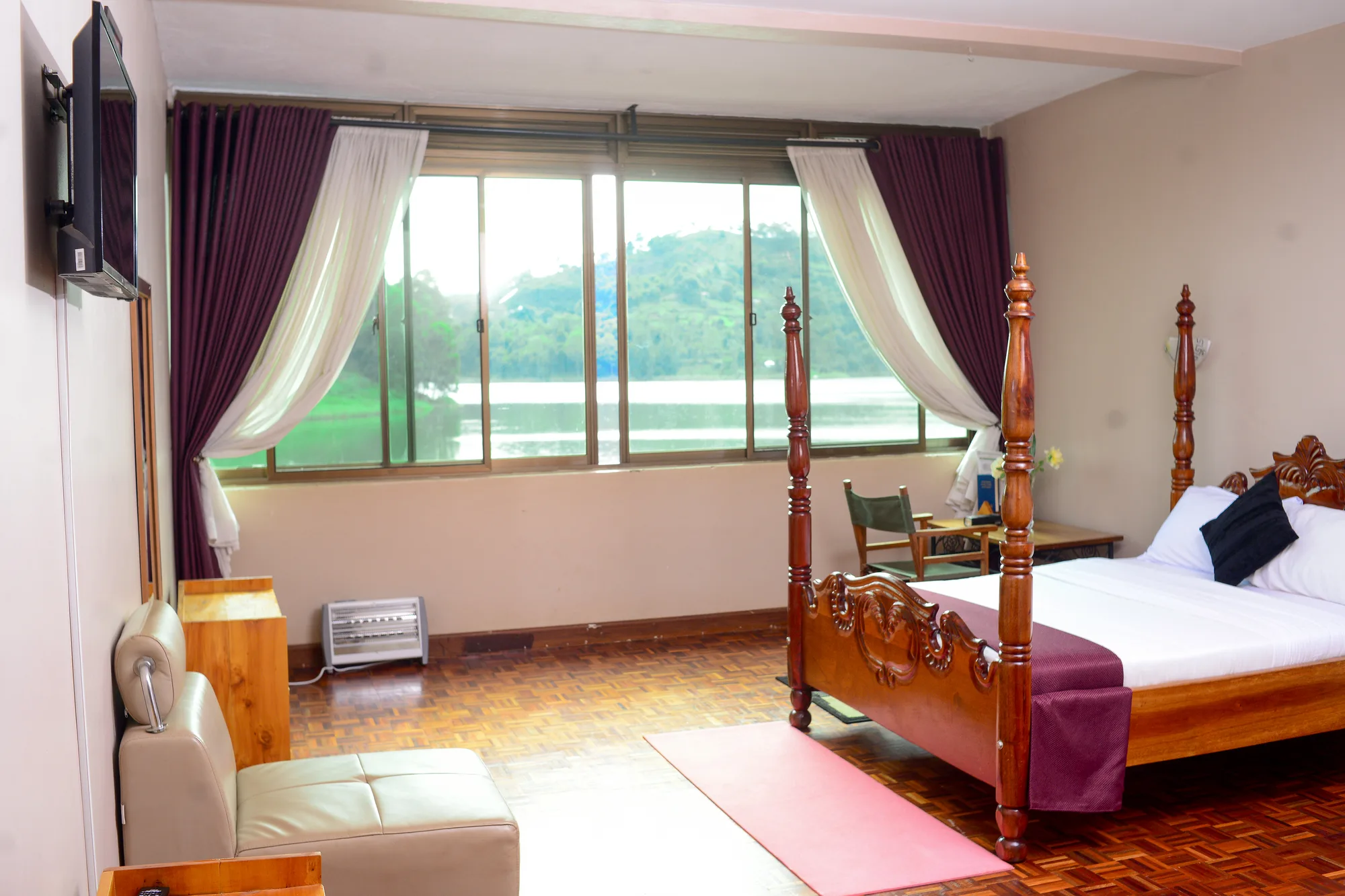 A guest room with a carved four-poster bed, sofa, wall-mounted television and a wide window looking onto Lake Bunyonyi