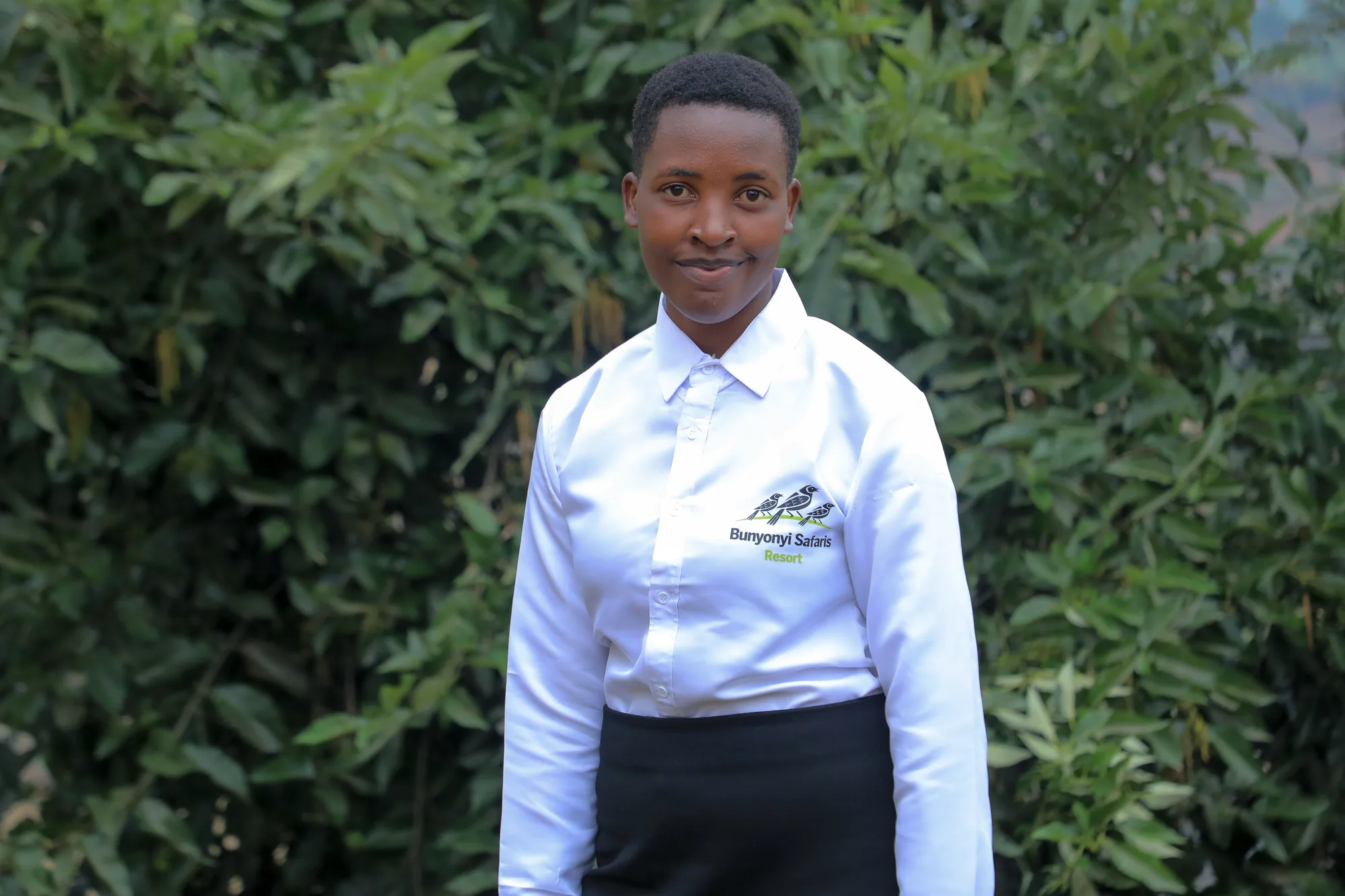 A staff member in a branded white shirt standing in front of green foliage